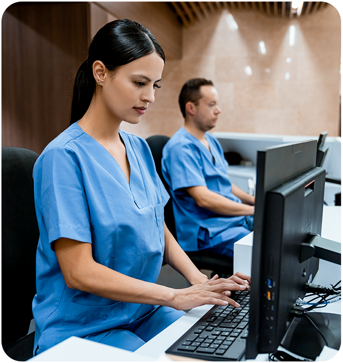 A healthcare professional wearing blue scrubs is seated at a desk, typing on a computer in a modern medical facility. Another professional in blue scrubs is seated in the background, also working at a computer. The setting is well-lit with a clean and professional appearance.