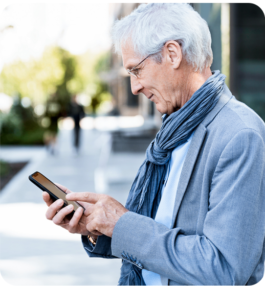 A doctor dressed casually in a collared shirt and blazer, without a white coat or stethoscope, is standing outdoors, looking at their smartphone. They appear focused as they type or scroll on the device. The background shows a blurred urban setting with greenery and sunlight.